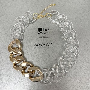 Puede incluir: Un collar de cadena de plástico transparente y dorado. El collar tiene una cadena dorada gruesa de un lado y una cadena de plástico transparente del otro. El collar está etiquetado como "Style 02".