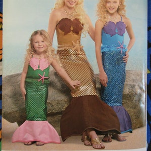 May include: Simplicity sewing pattern for mermaid costumes. The pattern features three mermaid costumes with seashell tops and scale-patterned skirts in gold, blue, and green. The pattern includes the text "U.S. ALL SIZES" and "WWW.ETSY.COM/SHOP/THEHALLOWEENTREE".