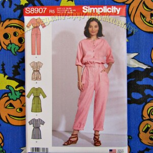 May include: Simplicity sewing pattern S8907 featuring a pink jumpsuit with a button-down top and belted waist. The pattern includes variations of jumpsuits and rompers. The model is wearing the jumpsuit. Size U.S. 14-22.