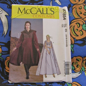May include: A McCall's Costumes sewing pattern, M7854, featuring illustrations of medieval-style gowns and capes. The pattern envelope is white and yellow, with the size E5 (14-16-18-20-22) printed on the side. The background is a blue fabric with Halloween-themed patterns.