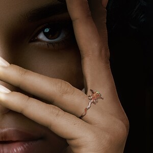 Puede incluir: Primer plano de una mano con un anillo de dise&ntilde;o floral. El anillo es de oro rosa y presenta una flor con peque&ntilde;as gemas brillantes. La banda del anillo tiene un dise&ntilde;o delicado y sinuoso.