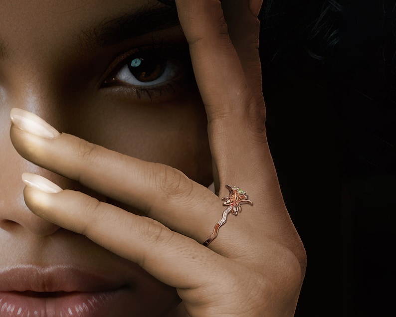 Puede incluir: Primer plano de una mano con un anillo de dise&ntilde;o floral. El anillo es de oro rosa y presenta una flor con peque&ntilde;as gemas brillantes. La banda del anillo tiene un dise&ntilde;o delicado y sinuoso.