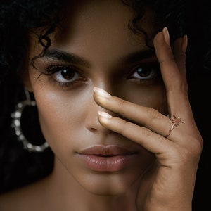 Puede incluir: Una mujer con cabello rizado oscuro y ojos marrones, usando un anillo de oro rosa con un dise&ntilde;o floral. El anillo est&aacute; en el dedo anular de su mano derecha, que cubre parcialmente su rostro. Tambi&eacute;n lleva grandes pendientes de aro plateados.