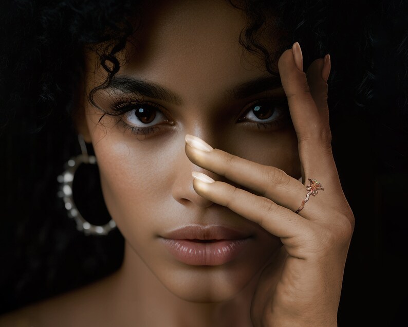 Puede incluir: Una mujer con cabello rizado oscuro y ojos marrones, usando un anillo de oro rosa con un dise&ntilde;o floral. El anillo est&aacute; en el dedo anular de su mano derecha, que cubre parcialmente su rostro. Tambi&eacute;n lleva grandes pendientes de aro plateados.