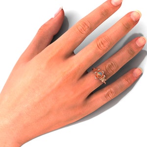 Puede incluir: Un anillo de oro rosa con una piedra preciosa de &oacute;palo, engastada en un dise&ntilde;o floral. El anillo est&aacute; en una mano sobre un fondo blanco. El &oacute;palo tiene una mezcla de colores, incluyendo blanco, naranja y azul.