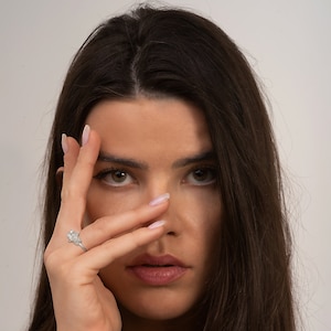 May include: A woman with long dark hair is looking at the camera. She is wearing a silver ring with a large diamond on her left ring finger. Her hand is covering part of her face.
