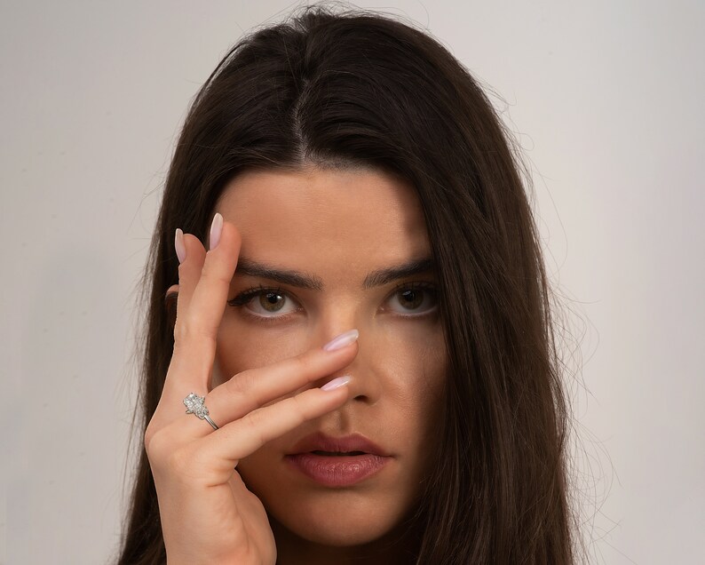 May include: A woman with long dark hair is looking at the camera. She is wearing a silver ring with a large diamond on her left ring finger. Her hand is covering part of her face.