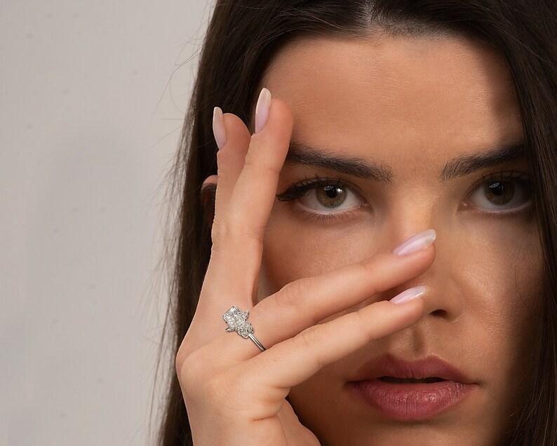 May include: A close-up of a woman's hand wearing a silver ring with a large, clear, square-shaped diamond. The ring has smaller diamonds on either side of the main stone.