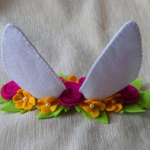 Puede incluir: Una diadema de fieltro blanco con dos orejas de conejo y una corona de flores. La corona est&aacute; hecha de flores de fieltro rosa y amarillo con centros de perlas blancas.