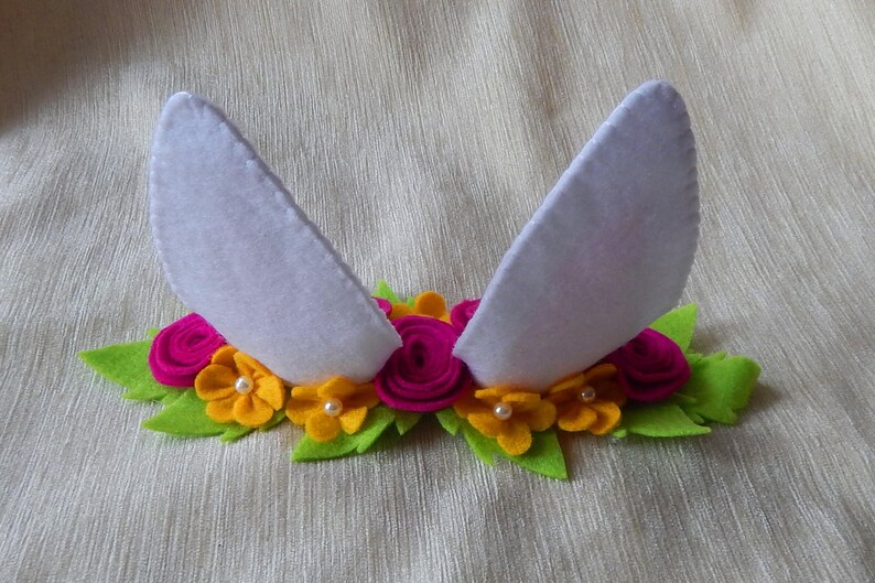 Puede incluir: Una diadema de fieltro blanco con dos orejas de conejo y una corona de flores. La corona est&aacute; hecha de flores de fieltro rosa y amarillo con centros de perlas blancas.
