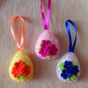 Op de afbeelding: Drie vilten paasei-ornamenten met linten. De eieren zijn geel, roze en wit, elk versierd met vilten bloemen in oranje, fuchsia en blauw, en versierd met kleine kralen. Een feestelijke paasdecoratie.