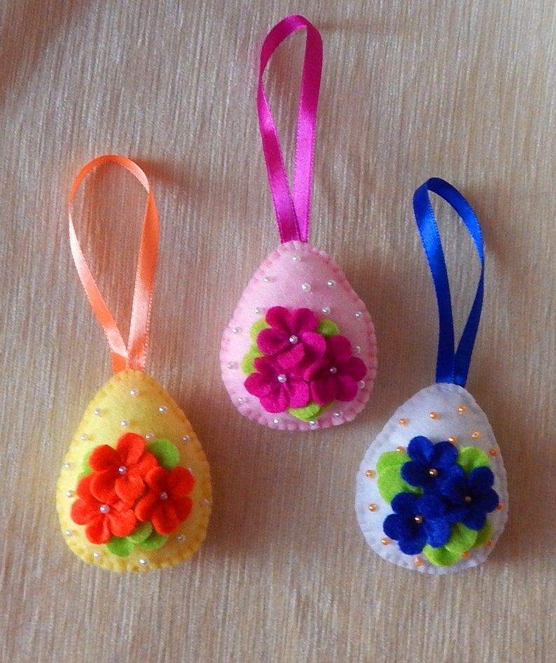Op de afbeelding: Drie vilten paasei-ornamenten met linten. De eieren zijn geel, roze en wit, elk versierd met vilten bloemen in oranje, fuchsia en blauw, en versierd met kleine kralen. Een feestelijke paasdecoratie.