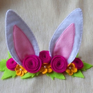 Puede incluir: Una diadema de fieltro con orejas de conejo rosas y hojas verdes con flores de fieltro rosas y amarillas.