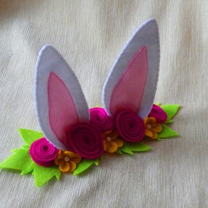 Puede incluir: Una diadema de orejas de conejo de fieltro con orejas rosas y una corona de flores de fieltro verde con flores rosas y amarillas.