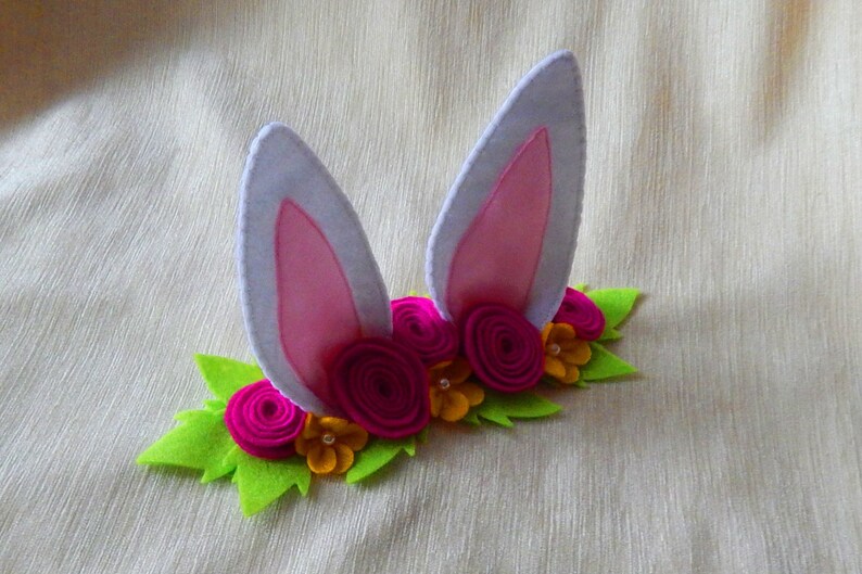 Puede incluir: Una diadema de orejas de conejo de fieltro con orejas rosas y una corona de flores de fieltro verde con flores rosas y amarillas.