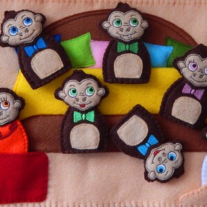 May include: A felt finger puppet play set featuring Seven brown monkeys with different colored bow ties. The monkeys are sitting on a yellow bed with brown and pink pillows. The set is perfect for imaginative play and storytelling.