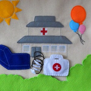Puede incluir: Un conjunto de juego de fieltro con un sill&oacute;n azul, un resorte, un botiqu&iacute;n blanco con una cruz roja, un edificio gris con una cruz roja, un sol amarillo y tres globos en naranja, azul y rosa.