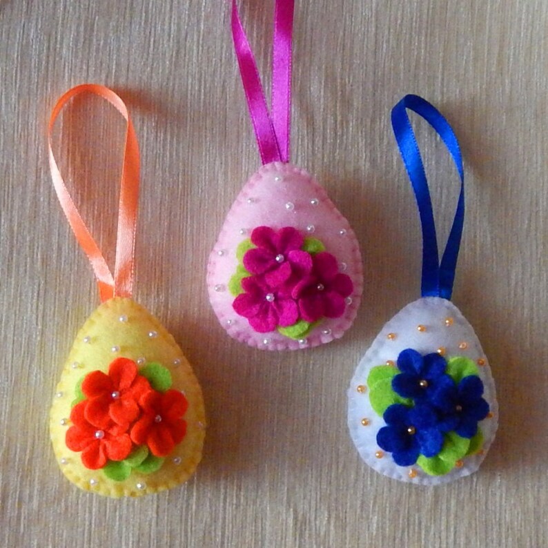 Op de afbeelding: Drie vilten paasei-ornamenten met linten om op te hangen. E&eacute;n is geel met oranje bloemen, een andere is roze met magenta bloemen en de laatste is wit met blauwe bloemen. Allemaal versierd met kleine kralen.