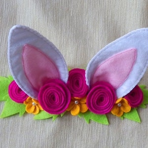 Puede incluir: Una diadema de fieltro con orejas de conejo rosas y un arreglo floral. Las flores est&aacute;n hechas de fieltro y son de color rosa, amarillo y verde.