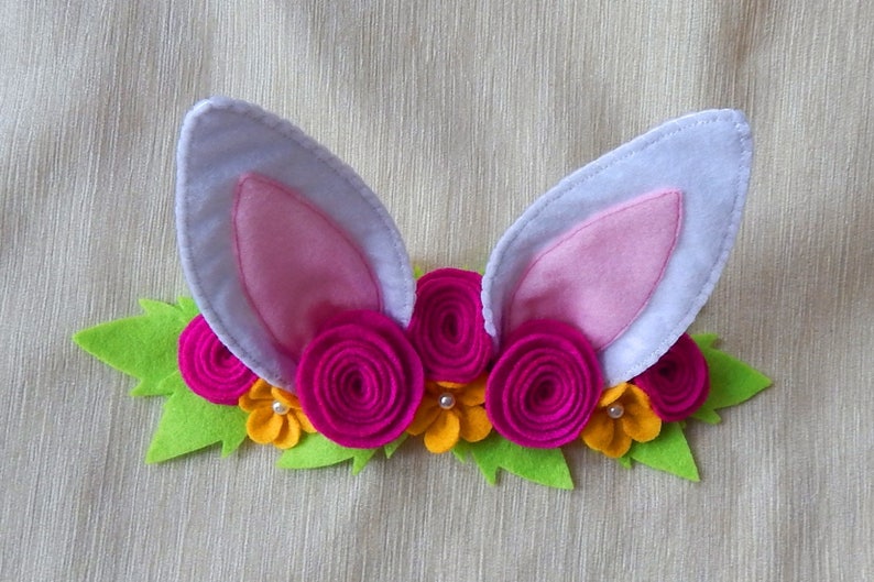 Puede incluir: Una diadema de fieltro con orejas de conejo rosas y un arreglo floral. Las flores est&aacute;n hechas de fieltro y son de color rosa, amarillo y verde.