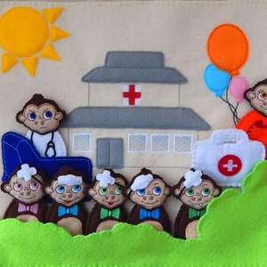 Puede incluir: Un juego de fieltro con un edificio de hospital, un sol, un botiqu&iacute;n de primeros auxilios y seis monos. Un mono es un m&eacute;dico con un estetoscopio y una silla azul. Los otros monos son pacientes con vendas en la cabeza. Los monos son marrones con caras blancas y corbatas de lazo azules. El edificio del hospital es gris con una cruz roja en la parte delantera. El sol es amarillo con rayos naranjas. El botiqu&iacute;n de primeros auxilios es blanco con una cruz roja en la parte delantera.
