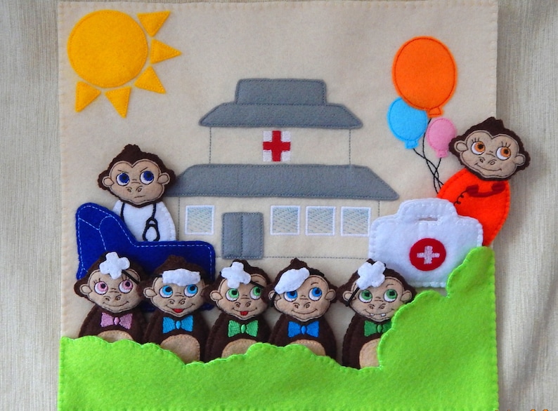 Puede incluir: Un juego de fieltro con un edificio de hospital, un sol, un botiqu&iacute;n de primeros auxilios y seis monos. Un mono es un m&eacute;dico con un estetoscopio y una silla azul. Los otros monos son pacientes con vendas en la cabeza. Los monos son marrones con caras blancas y corbatas de lazo azules. El edificio del hospital es gris con una cruz roja en la parte delantera. El sol es amarillo con rayos naranjas. El botiqu&iacute;n de primeros auxilios es blanco con una cruz roja en la parte delantera.