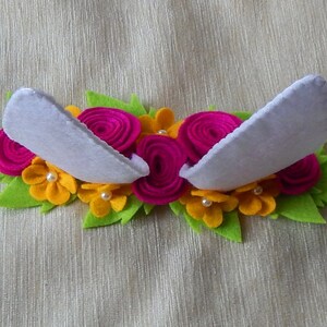 Puede incluir: Una diadema de fieltro blanco con dos orejas de fieltro blanco y un dise&ntilde;o floral. Las flores son de fieltro rosa y amarillo y tienen centros de perlas blancas.