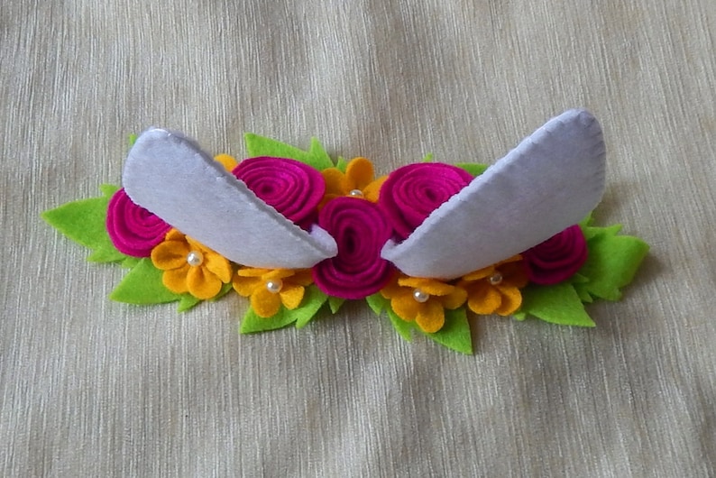 Puede incluir: Una diadema de fieltro blanco con dos orejas de fieltro blanco y un dise&ntilde;o floral. Las flores son de fieltro rosa y amarillo y tienen centros de perlas blancas.