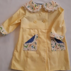 May include: A yellow child's coat with a white lace collar and floral print fabric. The coat has two pockets with blue and white floral fabric and white lace trim. The text on the fabric says "Forget me not".