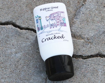 May include: A white plastic bottle of "Hippie Goat Lotion" with a black cap. The bottle has a cartoon illustration of a goat and the text "Cracked" on the front. The bottle is sitting on a cracked concrete surface.