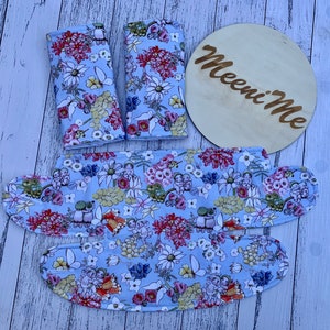 May include: A set of four blue fabric baby stroller straps with a floral print. The straps are decorated with a variety of flowers, including daisies, and have a light blue background. The straps are arranged on a white wooden surface. The straps are labeled "Meeni Me".