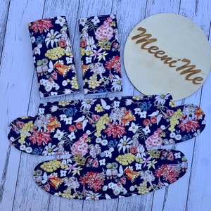 May include: A set of three fabric car seat protectors with a floral pattern. The protectors are navy blue with yellow, red, and white flowers. The fabric has a white border. The set includes two armrest covers and a headrest cover. The headrest cover is shaped like a crescent moon. The set is labeled "Meeni Me".
