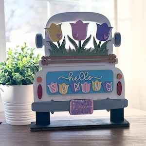 Puede incluir: Un camión decorativo de madera con carrocería blanca, detalles grises y base negra. La caja del camión presenta tres tulipanes coloridos y las palabras "hello SPRING". La matrícula dice "Flower Market".