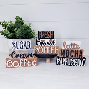 Puede incluir: Carteles de madera para una barra de café con las palabras "Sugar", "Cream", "Coffee", "Fresh", "Brewed", "Coffee", "Latte", "Mocha" y "Cappuccino". Los carteles son de varios colores y fuentes, con una planta en una maceta blanca.