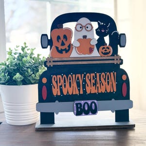 Könnte beinhalten: Eine schwarze Holz-LKW-Dekoration mit den Worten "Spooky Season" in Orange. Die Ladefläche des LKWs enthält einen Kürbis, einen Geist mit Brille, der einen Kürbis hält, eine schwarze Katze und einen weiteren Kürbis. Auf der Stoßstange steht "BOO".