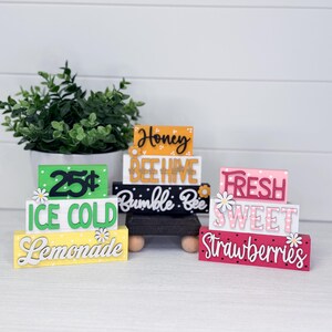 Könnte beinhalten: Eine Sammlung farbenfroher Holzschilder mit verschiedenen Aufschriften, darunter "Ice Cold Lemonade", "Honey Beehive" und "Fresh Sweet Strawberries". Jedes Schild ist mit einer Gänseblümchen-Akzent versehen und in leuchtenden Farben wie Gelb, Grün, Rosa und Schwarz bemalt.