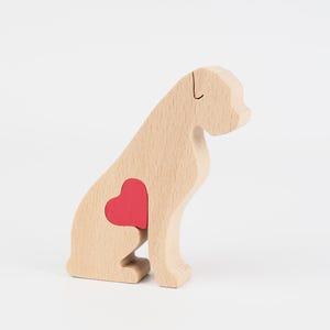 Puede incluir: Figura de perro de madera con una incrustación en forma de corazón rojo. El perro es de color marrón claro, con un diseño simple y estilizado. El corazón es de un rojo vibrante, contrastando con la veta natural de la madera. El perro está sentado.