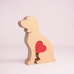 Op de afbeelding: Een houten hondenfiguur met een rode hartvormige uitsparing in zijn borst. Het figuur is gemaakt van lichtbruin hout en staat op een wit oppervlak.