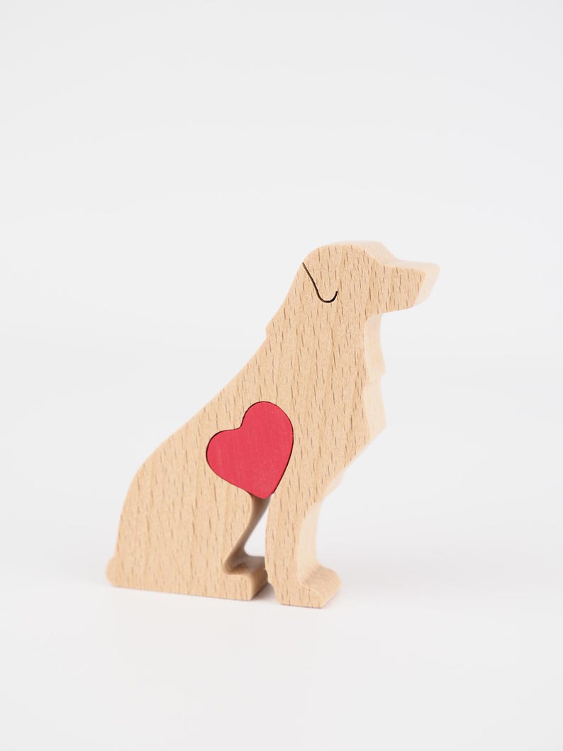 May include: A wooden dog figurine in a sitting position, featuring a red heart-shaped inlay on its side. The dog is made of light-colored wood with visible grain, and a black line defines the eye. The background is plain white.