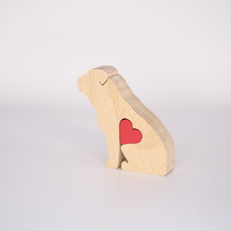 Puede incluir: Una figurita de perro de madera con un coraz&oacute;n rojo recortado en su pecho. La figurita est&aacute; hecha de madera clara y tiene un dise&ntilde;o simple y minimalista.