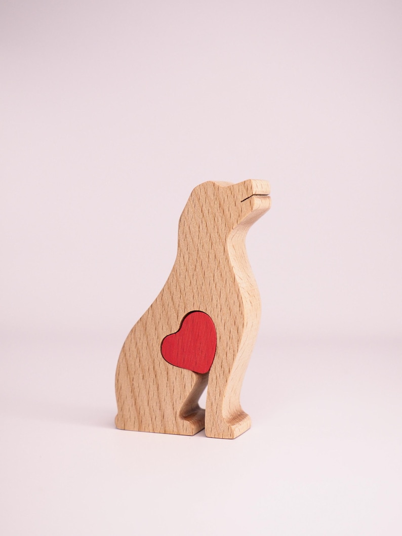 May include: Wooden dog figurine with a red heart cutout in its chest. The figurine is made of light brown wood and is sitting on a white surface.