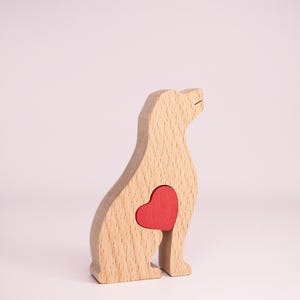 May include: Wooden dog figurine with a red heart cutout in the chest area. The figurine is made of light wood and is sitting on a white surface.