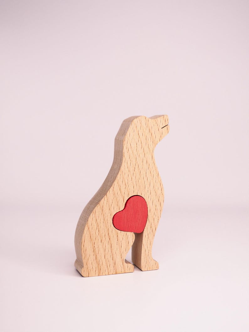 May include: Wooden dog figurine with a red heart cutout in the chest area. The figurine is made of light wood and is sitting on a white surface.