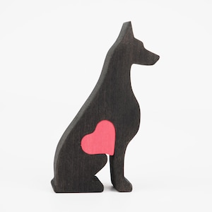 May include: A wooden silhouette of a sitting dog, painted black with a red heart-shaped cutout on its side. The dog has a pointed ear and a slightly raised tail. The background is plain white.