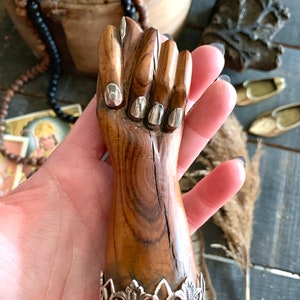 May include: A carved wooden fist figurine with silver-colored accents on the fingernails and a decorative silver base. The wood has a natural grain and a warm brown tone, creating a rustic aesthetic.
