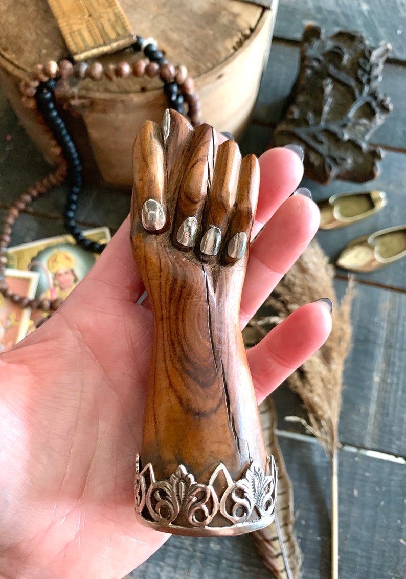 May include: A carved wooden fist figurine with silver-colored accents on the fingernails and a decorative silver base. The wood has a natural grain and a warm brown tone, creating a rustic aesthetic.