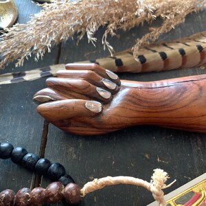 May include: A wooden hand sculpture with silver-painted fingernails. The sculpture is brown with visible wood grain. A strand of black beads and a feather are also visible.