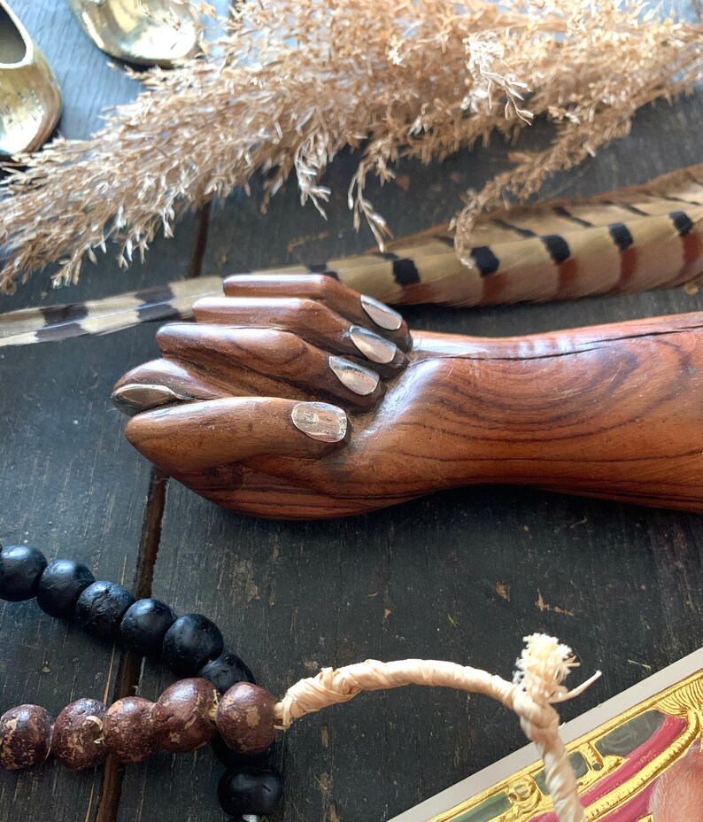 May include: A wooden hand sculpture with silver-painted fingernails. The sculpture is brown with visible wood grain. A strand of black beads and a feather are also visible.
