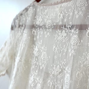 May include: Close-up of a sheer, white lace garment with floral and leaf patterns. The delicate fabric is draped over a wooden hanger. The neckline is trimmed with a scalloped edge.