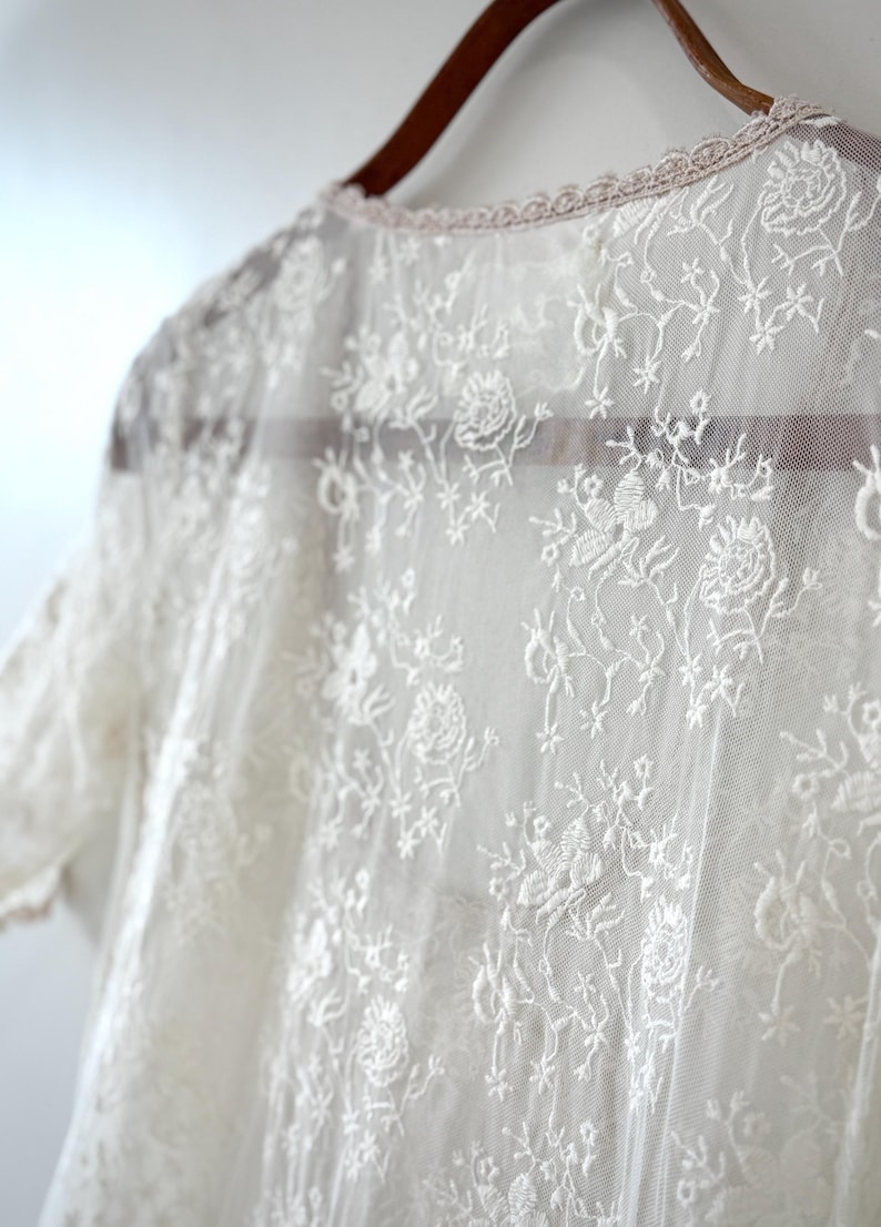 May include: Close-up of a sheer, white lace garment with floral and leaf patterns. The delicate fabric is draped over a wooden hanger. The neckline is trimmed with a scalloped edge.